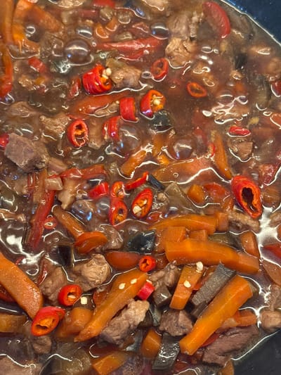 A close-up, overhead shot captures a rich, savory stew or braised dish simmering in a pot. The main subjects are various food ingredients immersed in a dark, glossy, and thick sauce.

Prominently featured are generous chunks of cooked meat, appearing tender and brown. Interspersed among the meat are numerous pieces of vibrant orange carrots, cut into irregular, elongated shapes. Adding a pop of color and likely heat to the dish are sliced red chili peppers, some appearing as rings with visible seeds, others as longer strips. There are also darker, almost blackish, irregular pieces that could be other vegetables like eggplant or mushrooms, or even additional meat or fat components.

The surface of the dark brown sauce reflects light, creating glossy highlights and indicating a thick consistency. The ingredients appear well-cooked, suggesting the dish has been simmering for some time, allowing flavors to meld. The overall impression is one of a hearty and flavorful meal.

No people are visible in the image, and there is no discernible text. The image provides no specific location cues beyond the context of being food, nor does it indicate the time of day or weather.