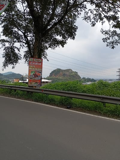 The image shows a roadside view of a paved road in Kutawaringin, Indonesia. A large tree occupies the left side of the image, partially obscuring a sign for a restaurant serving "Mie Kocok Gawir Sate Maranggi." The sign is mounted on a pole and has a red background with white lettering. The road is bordered by a metal guardrail and a grassy verge. In the distance, a rocky hill covered in green foliage rises up on the right side. A power line runs across the top of the image. The sky is overcast. The coordinates provided suggest the location is on Jalan Raya Sampit - Pangkalan Bun, a main road running through Kutawaringin.