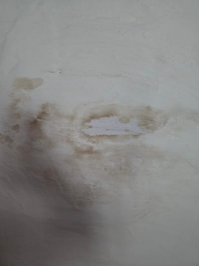 The image shows a close-up of a textured, off-white wall with visible water stains. The stains are a light to medium brown color and appear to be spreading across a section of the wall, with some areas showing lighter, more diluted discoloration and a central patch of lighter, possibly repaired or cleaner plaster. The texture of the wall is uneven, with subtle ridges and small imperfections. The lighting appears to be coming from the upper left, casting a slight shadow on the lower right portion of the image. There are no people, objects, or discernible actions depicted in this image. The scene is purely an abstract detail of a damaged wall surface.