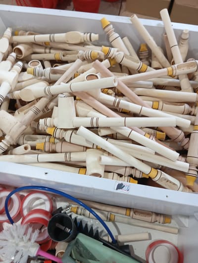 The image shows a bin filled with unfinished wooden smoking pipes. Some of the pipes have yellow accents or brown stripes. The bin is made of white plastic or metal. Below the bin are various cleaning supplies including brushes and rolls of tape. The image may have been taken in a retail or workshop setting in Eshanguzar, Uzbekistan, given the request.