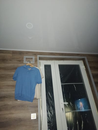 The image depicts an indoor scene with a wooden-paneled wall and a white ceiling. A blue polo shirt hangs on a hanger on the wall. To the right, a white door with a glass panel is visible, covered with black plastic sheeting. The door has a white handle and lock. A small square light fixture is mounted on the wooden wall. The scene is likely an interior of a residential or commercial building in Rudamina, Lithuania.