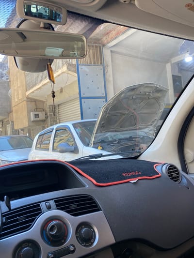 The image shows a view from inside a car, likely a Kangoo van, in Baraki, Algeria. The car's hood is open, and the view through the windshield shows a street with buildings, likely a repair shop or service area. The car's interior, including the dashboard with climate control knobs, is visible. The rearview mirror is also in the frame.