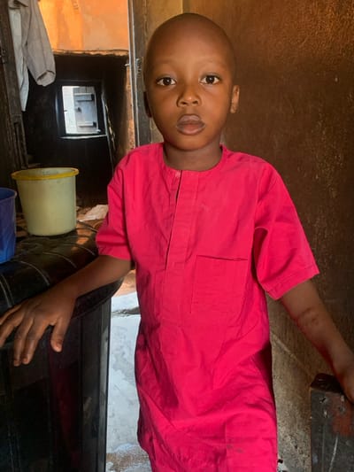 A young Black boy, approximately 4-7 years old, stands centered in what appears to be an indoor residential or communal space in Okene, Nigeria. He has a bald head and a neutral expression, looking directly into the camera with dark eyes and slightly parted lips. He wears a short-sleeved, bright pink tunic with a mandarin collar, a vertical placket, and a left breast pocket. His left hand rests on a dark, curved, possibly upholstered surface, while his right hand rests on a dark, angular, possibly wooden post or door frame.
In the background to the boy's left, a light-colored garment is hanging. Below this, a doorway opens into a darker interior, revealing a smaller, rectangular opening or window further back, through which vague structural elements are discernible. Two plastic buckets, one light yellow with a green rim and another dark blue, are visible on a surface in the lower-left background. The walls in the immediate background are rough-textured and exhibit earthy tones of brown and beige. The scene is illuminated by ambient light, with a brighter light source implied from the distant opening.