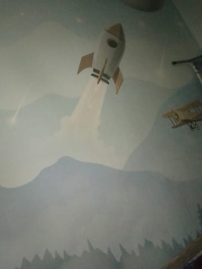 This image depicts a mural painted on a wall. The mural features a rocket ship ascending into the sky with smoke trailing behind it. To the right of the rocket, an older biplane is visible in flight. The background consists of stylized clouds and mountains. At the bottom of the mural, silhouetted pine trees suggest a forest. The overall color palette is muted blues and grays, with accents of white and tan for the vehicles. The mural is likely a decorative element within a building, potentially a child's room or a public space, and its style suggests a whimsical or imaginative theme. The location in Kalachinsk, Russia, is not evident from the visual content of the image alone.