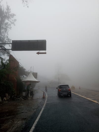 A car drives on a wet road through dense fog. A sign with a right-pointing arrow hangs above the road. A small business with an umbrella stands to the left. Two people are standing under the umbrella. A motorcyclist is also visible on the road ahead. Visibility is extremely limited due to the fog, obscuring background details like buildings or distant objects.