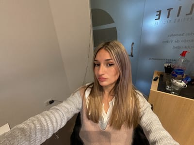 The image shows a young woman with blonde hair, wearing a white shirt and a light pink sweater vest, sitting in an office. She is looking directly at the camera. Behind her, there is a glass door with text on it, suggesting it might be a business or a professional space. On a wooden desk to her right, there are two glasses and a bottle of hand sanitizer. The overall lighting suggests an indoor environment, possibly an office in San Fernando del Valle de Catamarca, Argentina, given the context. The wall behind her is plain and light-colored, and there is an electrical outlet on the wall. The scene appears to be a candid or casual portrait taken in a workspace.