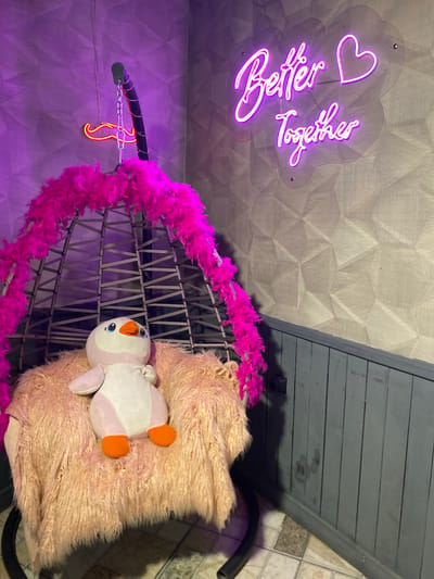 An indoor shot of a decorated corner in Kocasinan, Turkey, features a hanging chair with a plush pink feather boa and a fluffy pink blanket. A stuffed penguin sits in the chair. A neon sign that reads "Better Together" with a heart shape is mounted on the wall behind the chair, illuminated in purple. The wall has a geometric patterned wallpaper. To the left, a smaller neon mustache sign hangs from the chain supporting the chair, also illuminated in red. The floor is tiled in a mosaic pattern.
