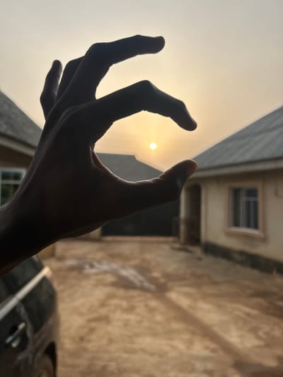 A silhouetted hand appears to "hold" the sun in an artistic photograph. The fingers are outstretched, forming a loose grasp around the glowing orb. The background features an urban or suburban landscape, including buildings with a window and a vehicle, likely a car, partially visible. The overall composition is dramatic, playing with light and shadow to create a striking visual effect. The image is slightly tilted, adding a dynamic feel.