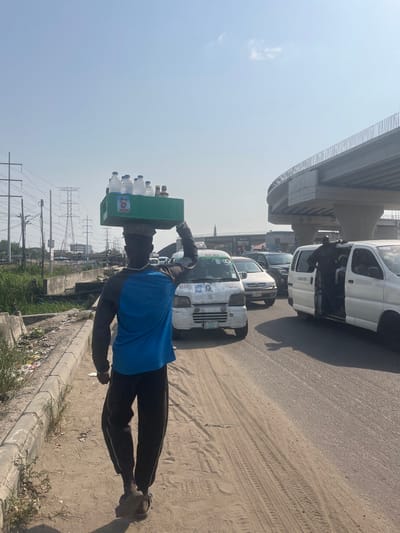The scene depicts a street in Ikota, Nigeria, under a clear, bright sky. In the foreground, an individual, seen from behind, walks along a sandy dirt shoulder adjacent to a paved road. The person wears a blue and black long-sleeved shirt, dark pants, and sandals, and carries a green plastic crate on their head, containing multiple plastic water bottles and several smaller brown bottles. A small poster depicting a man's face is affixed to the front of the crate. The person's left arm is raised and bent at the elbow, hand near the crate.
To the left of the walking individual, the dirt shoulder is bordered by a concrete curb, beyond which lies an unkempt strip of ground with sparse vegetation and litter, leading to an area with electrical poles and large transmission towers in the distant background.
To the right, on the paved road, traffic is present. Immediately behind the foreground person, a white compact van, likely a commercial passenger vehicle, is visible, featuring text that includes "BEKE" and two small posters of a man's face on its side. Further down the road, another white van has its side door open, with a person standing at the entrance. Additional vehicles are observed in various lanes. Overhead, a concrete elevated road or overpass structure with railings curves across the upper right portion of the image. In the mid-ground, a slender, dark, spire-like structure rises from beyond the traffic. The overall lighting indicates strong sunlight, casting visible shadows.