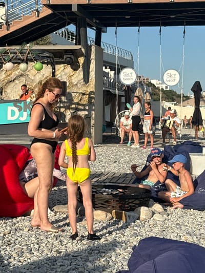 The image depicts a lively scene at what appears to be a beach club or resort, possibly in Yalta, Ukraine, given the provided context. Several people are visible, enjoying a sunny day outdoors. In the foreground, two women are seated on red beanbag chairs. One woman, wearing a black bikini, is looking towards the left, and the other, a young girl in a yellow bikini, is facing away from the camera. Behind them, more people are scattered on a gravelly surface, some reclining on blue blankets.
Further in the background, a row of white signs with circular logos and text that reads "Sky beach" are suspended from wires. Beyond these signs, buildings and lush green hills are visible, suggesting a coastal location with mountainous terrain. The overall atmosphere seems relaxed and casual, consistent with a vacation setting.
The time of day appears to be afternoon, indicated by the strong sunlight and shadows cast by the people and structures. The weather is clear and sunny, perfect for outdoor activities. The presence of beachwear and the "Sky beach" signs strongly point to a beachside or poolside establishment.
A metal fire pit is visible in the middle ground, adding an element of ambiance. The ground is covered in a mixture of gravel and paving stones. Among the people, some are dressed in swimwear, while others wear casual summer clothing. The overall impression is one of leisure and enjoyment on a warm day.