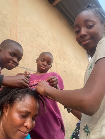 In Idiroko, Nigeria, an adult female is seated in the foreground, her head tilted forward and slightly to the left, with her eyes mostly closed and a faint smile. Her dark hair is in the process of being braided. Three children are positioned behind and above her, actively engaged in braiding her hair. To the left, a child with short dark hair, wearing a dark top, smiles broadly towards the viewer while braiding. In the center, another child with short dark hair, wearing a purple t-shirt with partial text "MAKE KINDNESS...", focuses downwards on the braiding activity. To the right, a young female with dark braided hair, wearing a light-colored t-shirt with a tree pattern and a dark beaded bracelet on her right wrist, looks towards the camera with a slight smile while also braiding. The background consists of a light brown, textured wall, with a dark corrugated metal roof section visible in the upper right corner.