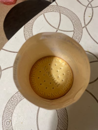 A round, golden-brown biscuit with an embossed pattern is visible inside a cylindrical, beige paper container. The biscuit is positioned at the bottom of the container, which is itself placed on a surface with a light-colored patterned tablecloth. The pattern on the tablecloth consists of interlocking circles and floral designs in shades of gray and beige. The paper container's rim is slightly curved inward, obscuring some of the upper edge of the biscuit. The embossed text on the biscuit is in a Cyrillic script and appears to read "КРЕМЕНЧУГСКОЕ" and "МАРИЙСКОЕ" which is a brand of cookies often found in Russia. There are no people or animals present in the image. The lighting suggests an indoor setting with diffused light. The specific location in Laishevo, Russia is not determinable from the visual information provided.