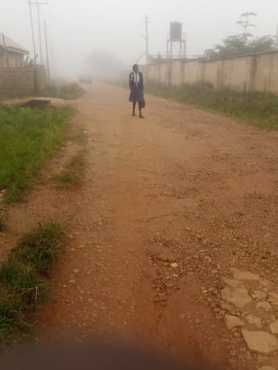 A lone individual, likely a student due to their uniform of a blue skirt, white shirt, and red tie, walks down a dirt road shrouded in dense fog in Katon Rikkos, Nigeria. The individual is holding a dark bag in their right hand and appears to be looking ahead. To the left of the road, a low brick wall borders a grassy area. Further down the road, a car is faintly visible through the fog. On the right side of the road, a tall concrete wall stretches into the distance, topped by a water tank on a metal structure. The ground is a mixture of red dirt and loose stones, with some patches of grass growing along the edges of the road. The overall atmosphere is obscured by heavy fog, limiting visibility and creating a somewhat eerie, muted environment.