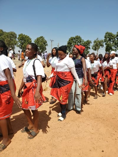 An outdoor scene in Bar Gada, Nigeria, depicts a procession of approximately 15-20 individuals, primarily young adult females, moving from left to right along a reddish-brown dirt path. The sky overhead is clear and light blue, indicating daylight. Numerous green-leafed trees line the background.
The participants are mostly attired in white short-sleeved tops and red wraps or skirts patterned with black and yellow/gold motifs, potentially depicting stylized birds. Several individuals wear green lanyards or ribbons around their necks. Footwear observed includes various flip-flops and sandals. One female near the center wears light blue ripped jeans underneath her red wrap, paired with black and white open-toed shoes. Another woman wears a dark blue top and a red patterned headwrap. Distinct shadows are cast on the ground, indicating direct sunlight. In the background, a utility pole with wires is visible, and additional people are present, suggesting a larger gathering. The overall appearance is that of a coordinated public event or cultural procession.