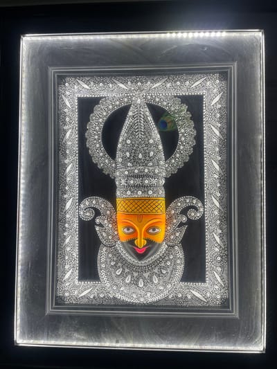 The image shows a framed artwork of a deity's face, possibly Hindu, rendered in a traditional Indian style. The deity has an orange face with large, expressive eyes and bright pink lips. The headdress and surrounding decorative elements are intricately detailed with white bead-like embellishments and fine line work against a black background. A single peacock feather is subtly placed to the right of the headdress. The artwork is framed in a silver-colored frame with LED lights illuminating the edges, creating a halo effect. The overall presentation suggests a devotional or artistic representation. There are no discernible elements that would place this image specifically in Ahmedabad, India, or any other particular city.