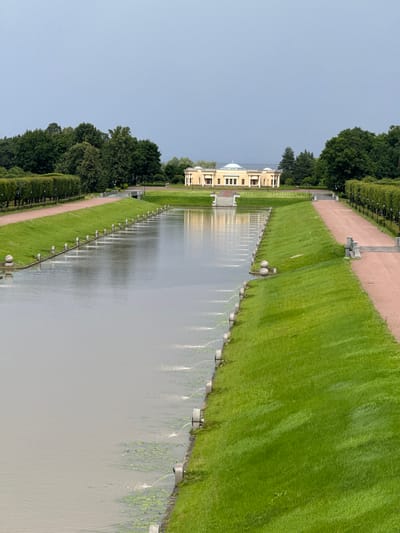 The image depicts a symmetrical formal garden with a long, straight canal. On the left side of the canal, manicured hedges line a pathway. On the right side of the canal, a grassy slope leads up to another pathway, also lined with hedges. Small fountains intermittently spray water into the canal from its right bank. In the background, at the far end of the canal, stands a pale yellow neoclassical building with white columns and a domed roof. The sky is overcast. This scene is characteristic of the Constantine Palace complex in Strelna, Russia, a suburban imperial residence located on the Gulf of Finland.
