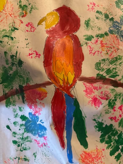 The image is a child's painting of a parrot perched on a tree branch amidst foliage and flowers. The parrot is rendered in red, orange, and yellow, with blue and green accents on its tail. The tree branch is brown. The surrounding foliage and flowers are depicted in various shades of green, pink, and blue using a stamping technique. The background is white. There is no location of Mèo Vạc Commune, Vietnam evident in the image.