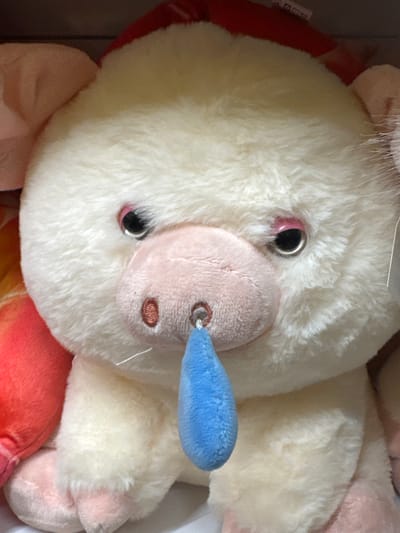 A close-up, eye-level shot shows a white, plush pig toy. The toy has pink ears, pink eyelids, and black, shiny eyes. Its snout is also pink, with two embroidered brown nostrils and a blue, plush object hanging from a metal ring in the center of the snout, resembling a drip or a pacifier. The background is out of focus and appears to be a shelf in a store. To the left of the pig, a corner of a red and orange plush toy is visible. The lighting is bright, suggesting an indoor setting. The image does not contain any information about the city of Ivanivka, Ukraine.