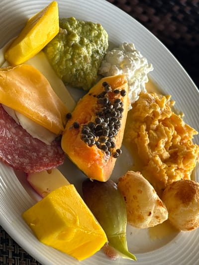 This close-up image showcases a diverse and appetizing breakfast or brunch spread laid out on a white, oval-shaped plate with a subtle ridged rim. No people are visible in the scene.
The plate is filled with a variety of food items. Prominently featured are fluffy, yellow scrambled eggs, appearing moist and freshly prepared, in the upper central portion. To the right of the eggs are two round, golden-brown baked items, which strongly resemble Brazilian Pão de Queijo (cheese bread). A vibrant orange half-slice of ripe papaya, replete with numerous dark, glossy seeds, sits centrally towards the bottom.
Accompanying the main items are several fresh components: bright yellow chunks of ripe mango are positioned on both the left and right sides of the plate. A dollop of white, lumpy cottage cheese or similar dairy product is visible in the upper left, while a mound of green, textured paste, likely guacamole or mashed avocado, sits below it. A whole, greenish-purple fig can be seen on the right side.
Beneath the tropical fruits, slices of various cheeses (ranging from pale yellow to orange-yellow) are layered with slices of reddish-brown salami or other cured meat, adding a savory element to the spread.
The arrangement of food suggests a hearty and well-presented meal, likely enjoyed during the daytime, possibly morning or early afternoon, given the breakfast/brunch typical items and soft lighting. The background features a dark, woven texture, suggesting a placemat or table surface. The combination of Pão de Queijo and tropical fruits like papaya, mango, and fig strongly points to a location with a warm climate, consistent with the given context of Rio de Janeiro, Brazil. There is no visible text in the image.
