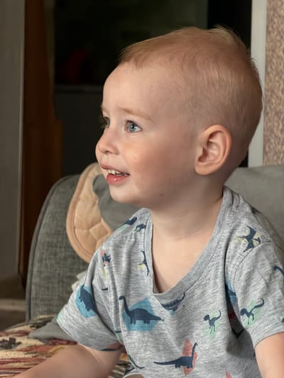 A close-up, sideways shot focuses on a young child with light blond hair and blue eyes. The child is wearing a gray t-shirt adorned with a pattern of blue and green dinosaurs. Their head is tilted, with their face looking to the left of the frame. The child's mouth is slightly open, revealing teeth and a hint of their tongue. The background is blurred, showing indistinct shapes and colors that suggest an indoor setting. A quilted tan object rests behind the child's neck, and a portion of a gray chair is visible. The lighting appears to be artificial, casting a warm glow on the child's skin. The overall mood is casual and intimate. No text is visible in the image.