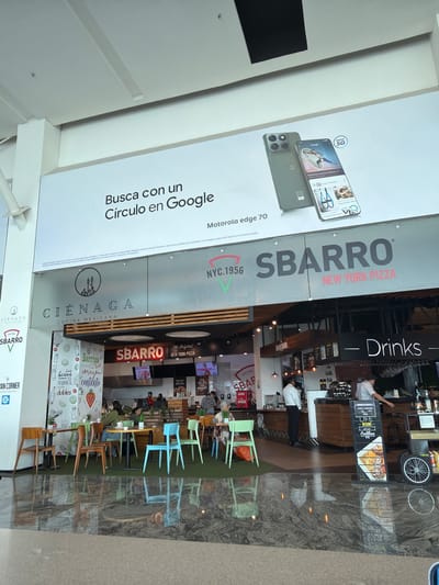 The image depicts a food court setting, potentially within a shopping mall or similar venue, located in Santa Cruz del Valle, Mexico. There are several food vendors: SBARRO New York Pizza, Cienaga cocina mexicana, and a Drink stand. A promotional advertisement for a Motorola edge 70 phone, highlighting the Google Lens feature, is posted on the left. There are tables and chairs are visible and a few people can be seen at the tables, indicating a relaxed, casual dining atmosphere. Reflections on the floor indicate that the food court is within a building with an entrance.