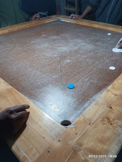 An overhead, angled view captures a wooden carrom board, dusted heavily with white powder, located in Maliat, Bangladesh. The board features a light brown playing surface with faint black grid lines and square corner pockets, one of which is visible in the foreground. A blue carrom piece (likely a striker) is positioned near the center-front, with three white carrom coins scattered on the right side of the board. Multiple individuals are present around the board, indicated by visible hands: one dark-skinned hand rests in the lower-left corner, another hand is on the upper-left frame, and a third hand is on the right side, with fingers touching a white carrom coin. Partial views of dark clothing are visible on the upper-left and upper-right sides. The background is dark and indistinct. The timestamp "02/12/2025 18:48" is visible in the bottom right corner.