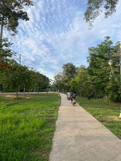 This is a medium shot, taken from a low-angle perspective, of a paved pathway in a park in Siem Reap, Cambodia. The pathway curves into the distance, bordered on the left by a lush green lawn and on the right by trees and a small canal. The sky is partly cloudy, with wispy white clouds against a blue background.
In the foreground, two people are riding motorbikes side-by-side on the pathway. The person in the front is wearing a white shirt with graphics and a helmet. The person behind is wearing a black shirt with graphics. Both motorbikes are white and have license plates.
The park is filled with a variety of trees, some with red flowers. There are also ornate streetlights along the pathway. The grass on the left is well-maintained, while the grass on the right appears a bit wilder. The water in the canal is murky brown.
The overall impression is one of a peaceful, natural setting within an urban environment. The lighting suggests it is either early morning or late afternoon, with the sun casting long shadows on the pathway.