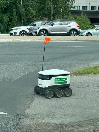 In Espoo, Finland, on a paved surface adjacent to a road, a white and green autonomous delivery robot with six wheels is positioned. The robot has "STARSHIP" branding and a green sticker that reads "KULJETAN KAUPAT RUOKAOSTOKSIA!" and a smaller graphic of a coffee cup. A black pole extends from the robot, topped with an orange triangular flag. In the background, a silver Volvo XC40 is parked, followed by a white Range Rover and a light blue sedan, all lined up on a roadside embankment. Green trees and a modern building with multiple windows are visible behind the vehicles. The road surface in the foreground shows varying textures of asphalt and gravel.