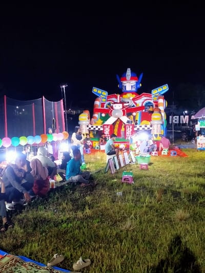 A large inflatable bouncy castle shaped like a robot stands in a grassy field under a night sky. The bouncy castle is brightly colored with red, blue, yellow, and white. There are several people sitting on the ground around the bouncy castle. They are all dressed in casual clothing. There are also some food stalls set up near the bouncy castle. The scene is illuminated by several lights. The coordinates provided indicate a location in a rural area, and the presence of food stalls and the bouncy castle suggests that this is a local event or fair.