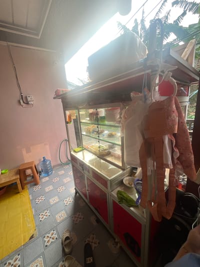 A street food stall is set up under a covered patio area. The stall features a glass display case filled with various food items, likely ingredients for dishes being prepared. Below the display case are red cabinets with black designs. Several items are hanging from the top of the stall, including cloth bags and what appears to be kitchen utensils. To the left of the stall, there is a pink wall with an electrical meter mounted on it. A large water dispenser sits on the floor next to a small orange stool. The floor is tiled with a patterned design. In the foreground, there are several pairs of footwear scattered around. The background shows lush greenery and trees, suggesting an outdoor or semi-outdoor setting, consistent with a location in Serdang Kulon, Indonesia. The lighting suggests it is daytime, with sunlight streaming in from the right side of the frame.