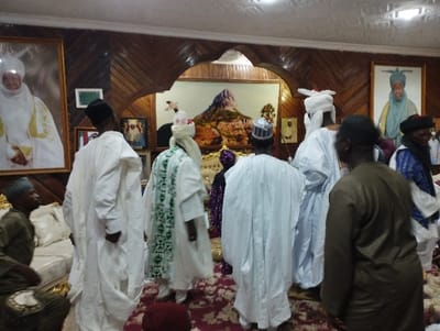 The image depicts a gathering of men in a room with wood-paneled walls and a carpeted floor in Kaltungo, Nigeria (latitude: 9.816, longitude: 11.315). There are nine men present, all wearing traditional white robes, except for one man in brown. They are standing and talking amongst themselves. There are several framed portraits on the walls. One portrait is of a man in white robes with a white turban. Another portrait shows a woman in white robes with a head covering. In the background is a large painting depicting a mountain range. The men are all standing in a relaxed, social setting. The room appears to be a living room or reception area.  The lighting is bright and the room is well-kept and clean.