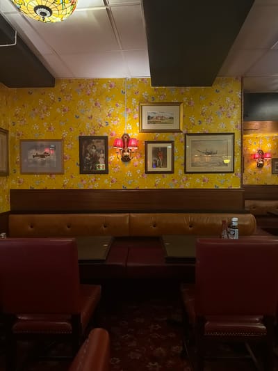 This is a photograph taken indoors, likely in a restaurant, featuring a wall with yellow floral wallpaper. Several framed pictures are displayed on the wallpaper, depicting various subjects including a landscape with a building, people in historical attire, and airplanes. Two red sconce lights with bell-shaped shades are mounted on the wall. Below the wallpaper is a wooden wainscoting. The seating consists of brown leather booths with button tufting, and in the foreground, there are red upholstered chairs. There are tables in front of the booths, and on one of the tables, a bottle of HP sauce and a small red bottle, possibly ketchup, are visible. The ceiling is paneled with recessed lights and a decorative stained-glass light fixture is visible in the upper left corner. The flooring is a patterned carpet. The image does not provide any information about the city of Lund, Sweden.