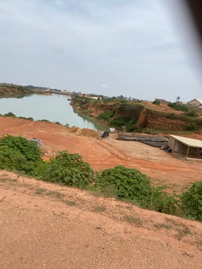 An aerial, slightly blurred view looks down upon a body of tranquil, light blue water, possibly a quarry lake, bordered by steep, reddish-brown earthen cliffs. The cliffs are sparsely covered with green vegetation, and the top of the cliffs are dotted with buildings. In the foreground, the landscape is a barren, reddish-orange dirt expanse, with mounds of dirt and piles of grey blocks. Several green bushes with broad leaves dot the foreground. In the middle ground, a cluster of grey blocks is arranged in rows, and a small, light-colored building with a slanted roof stands nearby. A white truck is parked to the left of the blocks. In the distance, a tall, slender tower pierces the overcast, grey sky. The overall scene suggests a construction or excavation site near a body of water, with residential buildings visible in the background.