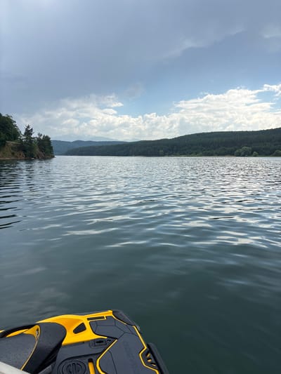 The image depicts a person's perspective from a yellow and black jet ski on a calm lake. The lake's surface is rippled, reflecting the sunlight and the overcast sky. A long, tree-covered hill or mountain range stretches across the background, bordering the far side of the lake. The sky is a mixture of blue and white clouds, with some areas appearing brighter than others, suggesting the sun is trying to break through.
On the right side of the frame, the rear section of the jet ski is visible, characterized by its black and yellow design. The jet ski is stationary, and the water around it is smooth. The scene appears to be set in a natural, tranquil environment.
On the left side of the frame, the shore of the lake is lined with trees and vegetation, indicating a lush natural setting. The overall mood is peaceful and serene, possibly suggesting a recreational activity on a pleasant day. The time of day appears to be daytime, given the brightness of the sky.
There is text visible on the jet ski, but it is partially obscured and difficult to read.
