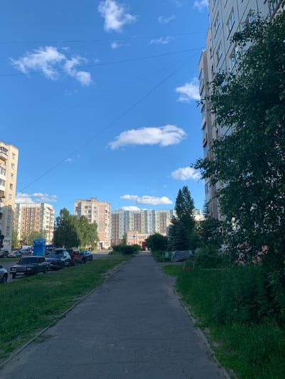The image depicts a street scene in Severodvinsk, Russia, under a clear blue sky with scattered white clouds. The perspective is slightly tilted, giving a dynamic feel to the urban environment. The foreground features a paved walkway bordering a strip of lush green grass, typical of a residential area. A row of parked cars lines the street, suggesting a daytime setting when people might be at work or out and about.
Behind the green strip, several multi-story apartment buildings are visible, showcasing a common architectural style with numerous windows. Some buildings appear older with a more muted color palette, while others are newer, displaying more vibrant colors like beige and light gray. Trees are interspersed between the buildings and along the street, adding a touch of nature to the urban landscape. In the distance, more apartment blocks punctuate the skyline.
The overall atmosphere is one of everyday life in a Russian city. There are no people clearly visible in the frame, and the focus is on the built environment and the natural elements that coexist within it. The sunlight suggests it is daytime, likely late morning or early afternoon, given the brightness and the shadows. The presence of parked cars and the general upkeep of the area indicate a functional and inhabited neighborhood. No discernible text is visible in the image.