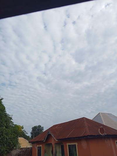 An outdoor shot captures a portion of buildings under a cloudy sky in Gindin Dutse, Nigeria. The foreground features the roof of an orange building with a rusty, corrugated metal roof. A satellite dish is attached to the side of this roof. To the left, a portion of a large, leafy green tree is visible. Behind the orange building, another building with a lighter colored, corrugated metal roof is partially visible. The sky is filled with scattered, textured clouds. Electrical wires are visible in the middle ground, stretching between utility poles.