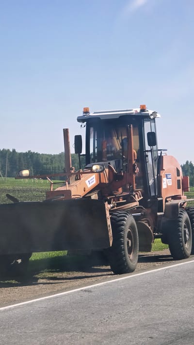 An orange road grader is parked on the side of an asphalt road. The grader is in the foreground, facing slightly to the right. It has a large metal blade at the front, large rubber tires, and a cab with windows. There are orange flashing lights on the roof of the cab. A person wearing an orange high-visibility vest is visible inside the cab. In the background, there is a green field with some vegetation and trees. The sky is clear and blue. The image appears to be taken during the daytime.