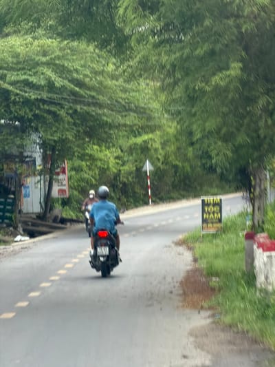 The image depicts a roadside scene in Dien Tho, Vietnam. A person on a dark-colored scooter is riding away from the camera on a paved road. The scooter has a red taillight illuminated. Another scooter, with two individuals on it, is visible further ahead on the road, also traveling in the same direction. The road is lined with lush green trees and vegetation. To the right of the road, a sign with yellow text on a black background reads "TIEM TOG NAM". A red and white striped pole is also present on the side of the road. On the left side of the road, there are some structures and what appear to be advertisements. The sky is overcast, suggesting a cloudy day.