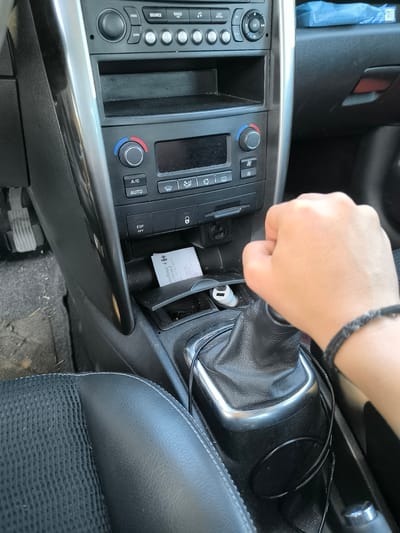 The image shows the interior of a car, likely a Peugeot 207 based on the dashboard layout. A hand is gripping the gear stick. The center console is visible, including the radio/CD player, climate controls, ESP off switch, and a small storage area containing a Carrefour supermarket receipt and a USB charger. The seating and interior trim are dark, and there's a black bracelet visible on the wrist of the hand gripping the gear stick. The location in Chaponnay, France, is not explicitly visible in the image but can be inferred if the Carrefour supermarket receipt has a Carrefour Contact location.