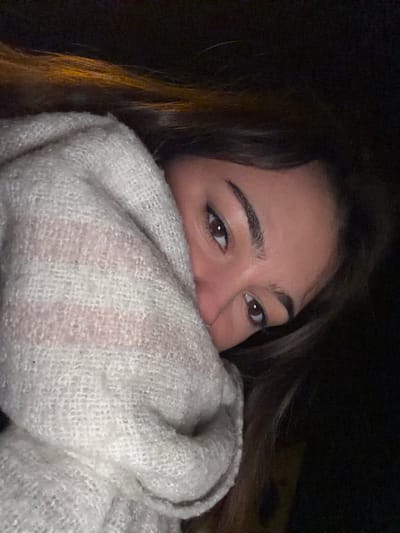 A young woman's face is partially obscured by a fluffy, off-white blanket or scarf. Her eyes, eyebrows, and nose are visible. She has brown eyes and hair, with a dark background. The lighting appears dim, creating a contrast between her face and the surrounding darkness.