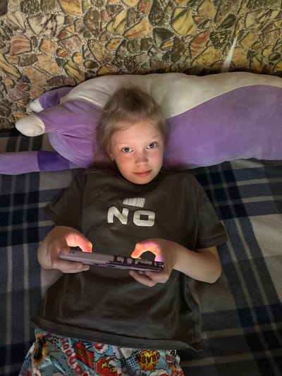 A young girl with blonde hair and blue eyes is lying on a plaid blanket, propped up against a large purple pillow. She is wearing a dark gray t-shirt with the word "NO" printed on it in white block letters, and colorful pajama bottoms adorned with comic book graphics. Her hands are holding a tablet, which is illuminated by its screen, casting a soft glow on her fingers. The background appears to be a textured wall with a stone-like pattern. The scene suggests a relaxed, indoor setting, possibly a bedroom or living room, in a residential area of Sayanogorsk, Russia.