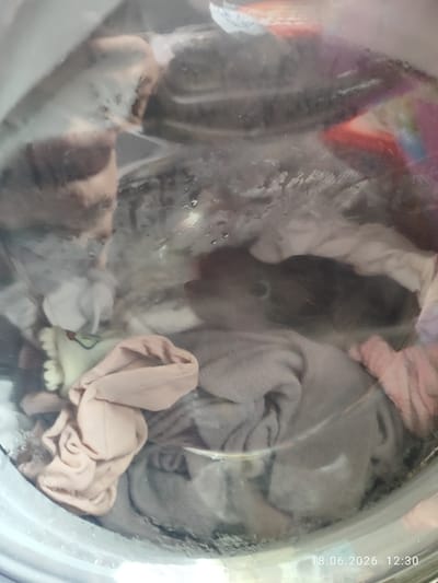 The image shows the inside of a washing machine drum filled with laundry. Several items of clothing, including towels and what appear to be t-shirts, are visible. Some are folded or bunched up, while others are submerged in water. The clothes are a mix of colors, including beige, grey, and pink, with some darker items also present. The washing machine drum is made of metal and glass. The image includes a timestamp in the bottom right corner: "18.06.2026 12:30". There is no indication of a specific location in Northampton, United Kingdom, within the image itself.