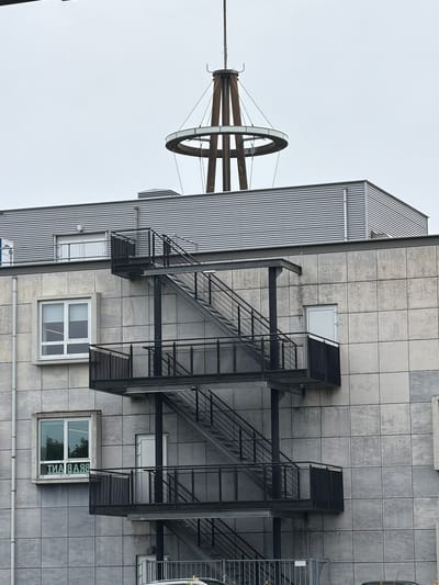 The image shows a modern building in the city of Son en Breugel, Netherlands. The building features a grey, tiled facade with a visible fire escape on the side. The fire escape is made of black metal and has multiple platforms and staircases. There are several windows on the building; one on the left side displays text in reverse, reading "BRAANT". At the top of the building, a corrugated metal roof is visible, and above that, a distinct, industrial-looking structure with a circular element and supporting beams. The sky is overcast. No people are visible in the image.