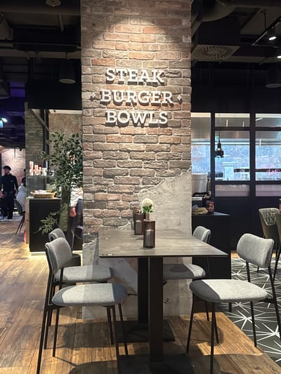 The interior shot shows a dining area, likely a restaurant. A brick wall with "STEAK BURGER BOWLS" vertically spelled out in white lettering is prominent. A long, grey tabletop is surrounded by modern grey chairs hanging from the ceiling. There are vases of flowers on the tabletop. Behind the brick wall, a service counter with decorative plants is visible. In the background, people are visible near the counter, suggesting a restaurant or eatery. The location of the restaurant is in Amstetten, Austria.
