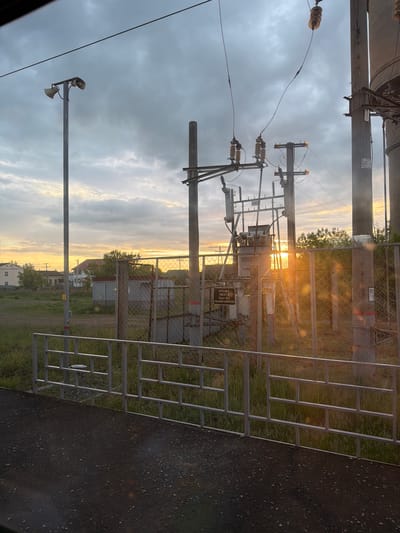The image depicts an outdoor scene in Kizner, Russia, captured near sunset. A chain-link fence with a metal frame is positioned prominently in the foreground, slightly obscuring the view. Behind the fence, an electrical substation is visible, featuring transformers, power poles, and various electrical components. A warning sign is attached to the substation's enclosure. The background shows houses and greenery, suggesting a residential or semi-rural area. The sky is overcast with clouds, tinged with the orange and yellow hues of the setting sun. The image appears to be taken from a moving vehicle, as evidenced by the blurry dark surface in the lower right corner, likely a road or pavement.
