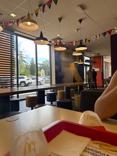 This image captures an interior view of a McDonald's restaurant in Zugdidi, Georgia. The scene is viewed from a table in the foreground, where a red tray holds what appears to be McDonald's food items, including a burger wrapper with the iconic golden arches logo. The restaurant's interior features numerous pendant lights with yellow and dark shades, suspended from a tiled ceiling. Decorative string flags of various national flags are hung along the ceiling.
Large windows line the back wall, offering a view of the exterior. Through the windows, several cars are visible parked outside, including white and silver SUVs and sedans. Beyond the parking lot, lush green trees and a bright sky suggest it is daytime. A small building with a blue roof and a statue on its facade is also visible in the distance, likely a religious building.
Inside the restaurant, several dark and yellow cylindrical stools are arranged around high-top tables. In the middle ground, two men are standing near a doorway. One man, wearing a gray polo shirt and dark pants, is looking down. The other man, in a black shirt and pants, is facing away from the camera. The restaurant walls are decorated with large black-and-white murals. An exit sign is visible above the doorway. The overall ambiance suggests a casual dining environment.