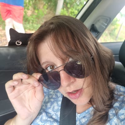 The image is a close-up selfie taken from inside a vehicle, likely a car. The perspective is from the passenger seat looking towards the driver's side. The woman in the foreground is wearing sunglasses pushed down her nose and is holding them with her left hand. She has brown hair, a light complexion, and is wearing a blue and white floral patterned shirt. Her lips are slightly parted, revealing teeth. In the background, out of focus, is another person wearing a blue shirt and red shorts, and the out-of-focus scenery suggests an outdoor environment with trees and foliage. The interior of the car is visible, including a seatbelt and a headrest. There is no specific indication of the location being Gangneung-si, South Korea.