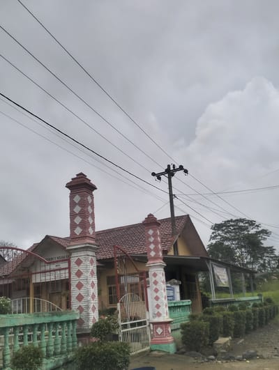 The image shows the entrance to a building, possibly a school, with a red-tiled roof and yellow walls. A decorative gate with red and white diamond patterns is visible, along with a green-painted fence. Overhead, multiple power lines stretch across a cloudy sky, supported by a utility pole. In the background, trees and vegetation are present. A sign above the entrance reads "SEKOLAH NEGERI 3 NISAM ANTARA". The overall scene depicts a building exterior in a possibly rural or suburban setting, with infrastructure elements such as power lines indicating an inhabited area. The image appears to be taken from a slightly elevated, ground-level perspective looking upwards towards the building and the sky.