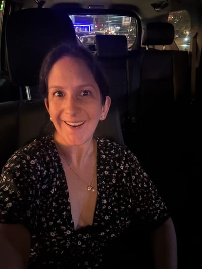 A woman is seated in the back seat of a car, possibly a taxi, at night. She is wearing a black dress with small white floral patterns and a gold necklace. She is smiling. Outside the car window, blurred lights and buildings can be seen, consistent with a street scene. The location is likely in Miraflores, Peru, based on visual landmarks.