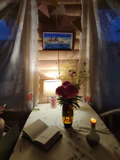 The image depicts a cozy indoor scene, likely a living room or dining area with a rustic wooden wall. A table is set with a floral-patterned tablecloth. On the table, an open book rests, its pages filled with text and illustrations. To the right, a lit candle in a decorative bottle flickers, casting a warm glow. A vase filled with pink and purple flowers stands near a lamp, which illuminates the scene. Above the table, a framed painting of a Venetian cityscape hangs on the wall, and colorful triangular flags are strung across the top of the room. Lace curtains adorn a window on the left, partially obscuring the dark blue sky outside. A person's hand, holding a phone, is visible on the far left of the frame, suggesting someone is present and engaged with their device. The overall ambiance is one of quiet relaxation and domestic comfort. There is no information available regarding the specific location within Бярэзінскі сельскі Савет, Belarus.